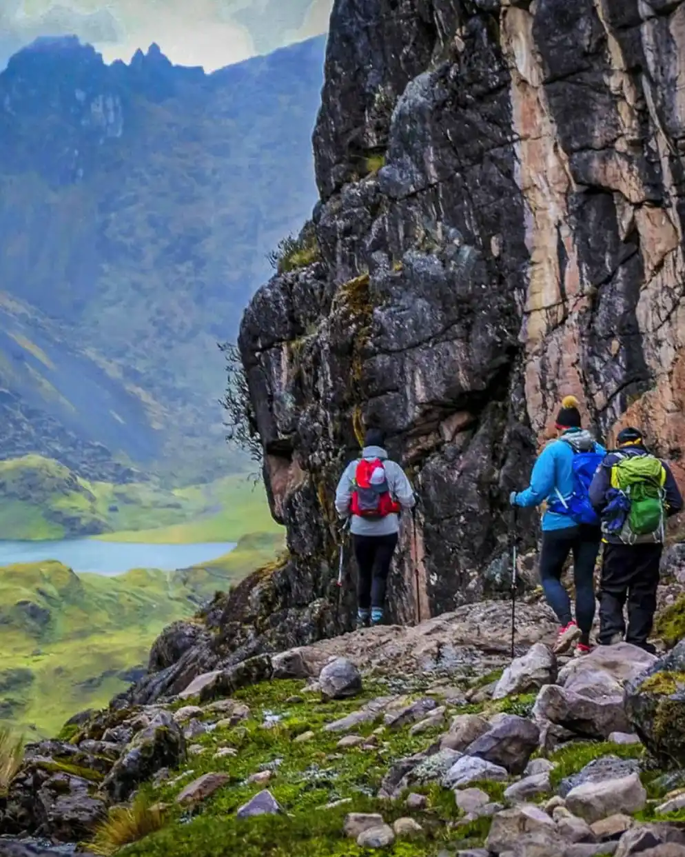 Grupo de excursionistas con mochilas y bastones camina junto a una pared rocosa en sendero de montaña en el camino de Lares trek.