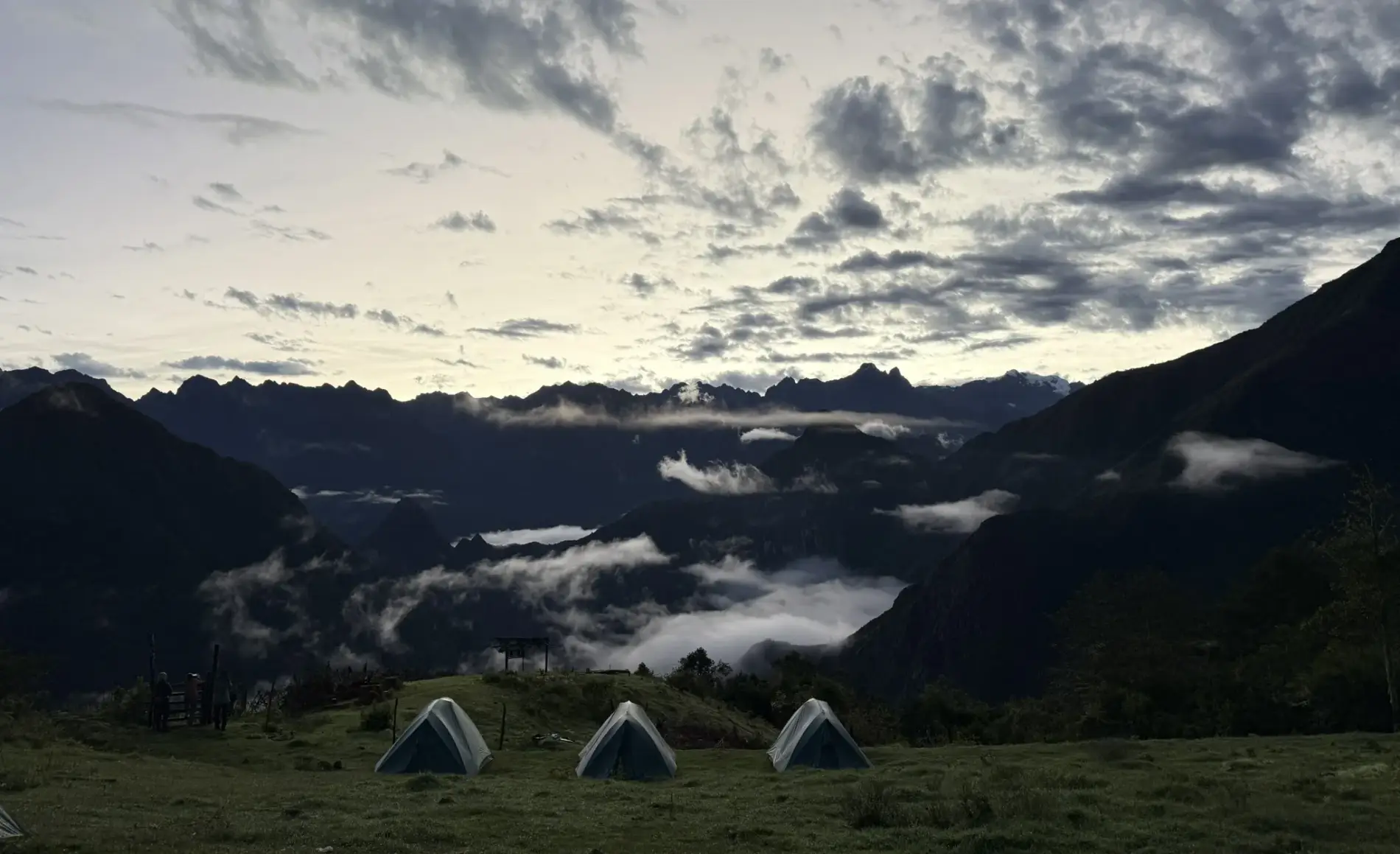 Series of tents set up at Peru Choquequirao trek campsite, surrounded by steep Andean mountains, grassy terraces and cloudy sky at dusk in Cusco, Peru.
