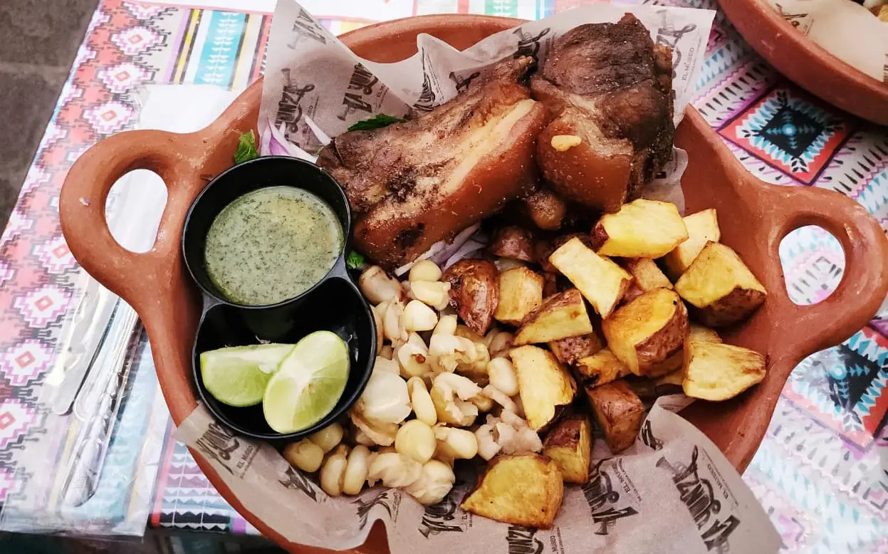 Clay platter with crispy pork chicharrón served with fried potato wedges, boiled corn, lime wedges, and green sauce on a patterned tablecloth.