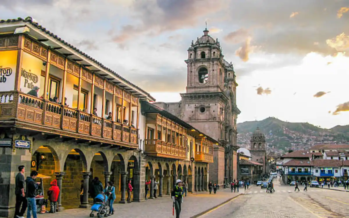 Calle histórica de Cusco con arcos coloniales, gente caminando y torre de iglesia bajo cielo dramático, arquitectura clásica del Perú.