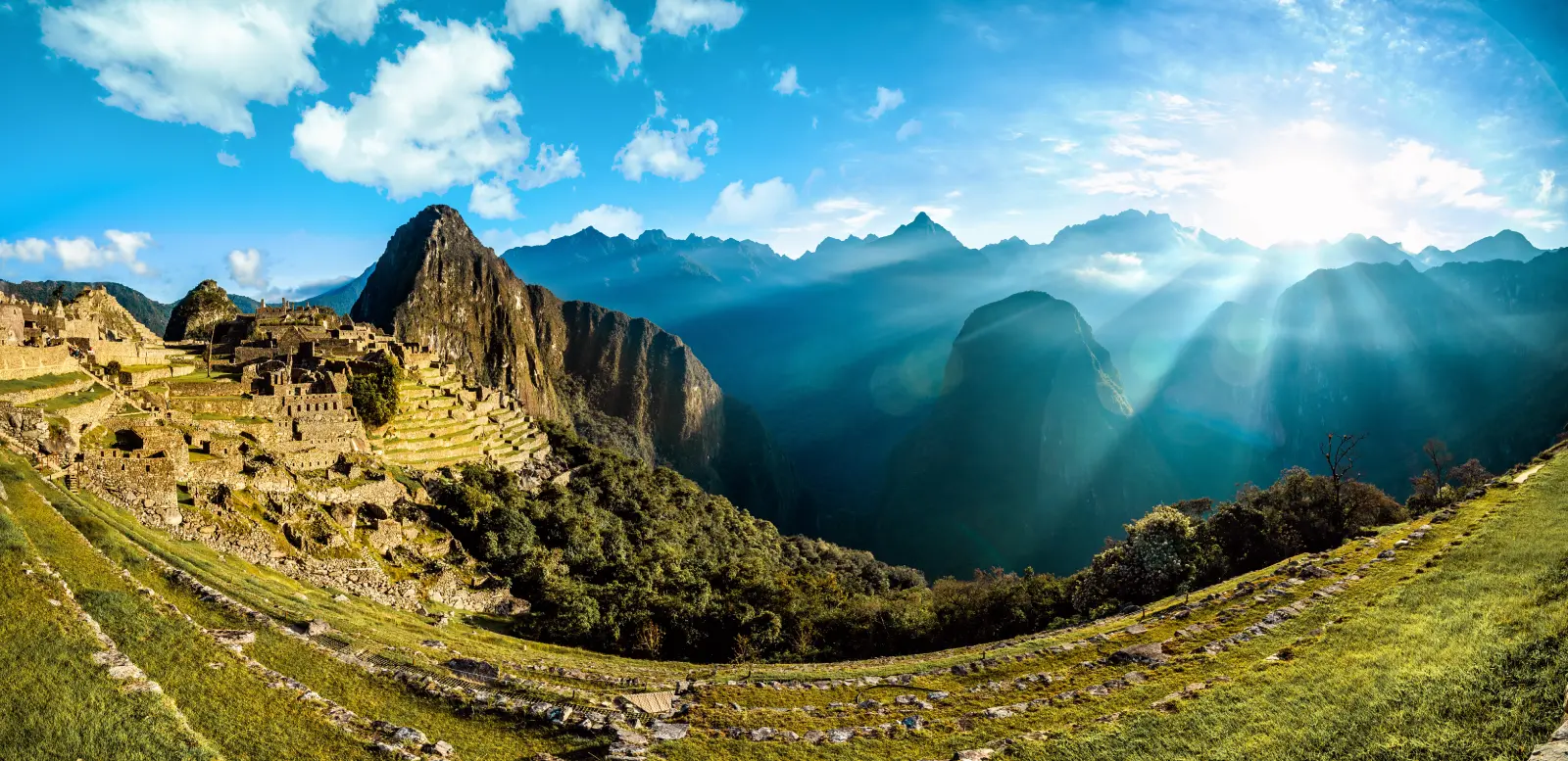 Panoramic sunrise view of Machu Picchu with blue sky, mountain silhouettes, and light spreading across the landscape