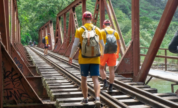 Two hikers with backpacks walking across a steel railway bridge toward Aguas Calientes on the Inca Jungle Trek to Machu Picchu.