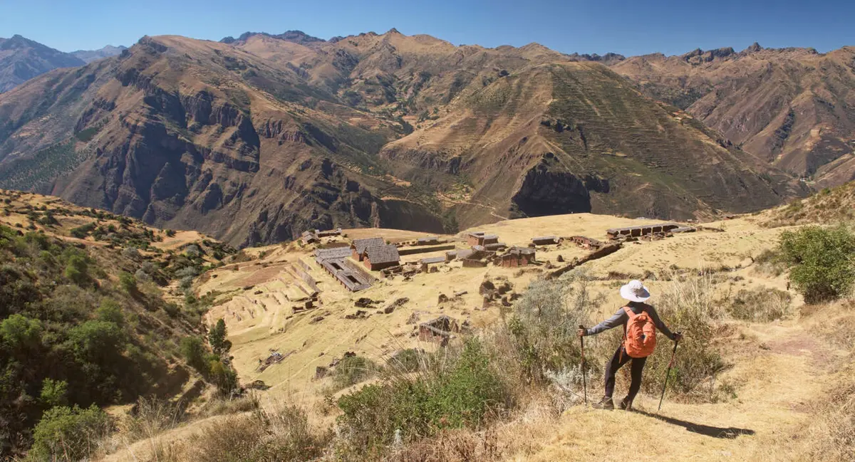 Hiker descending toward Huchuy Qosqo in the Sacred Valley, Cusco, with Inca terraces, dry hills and wide mountain views
