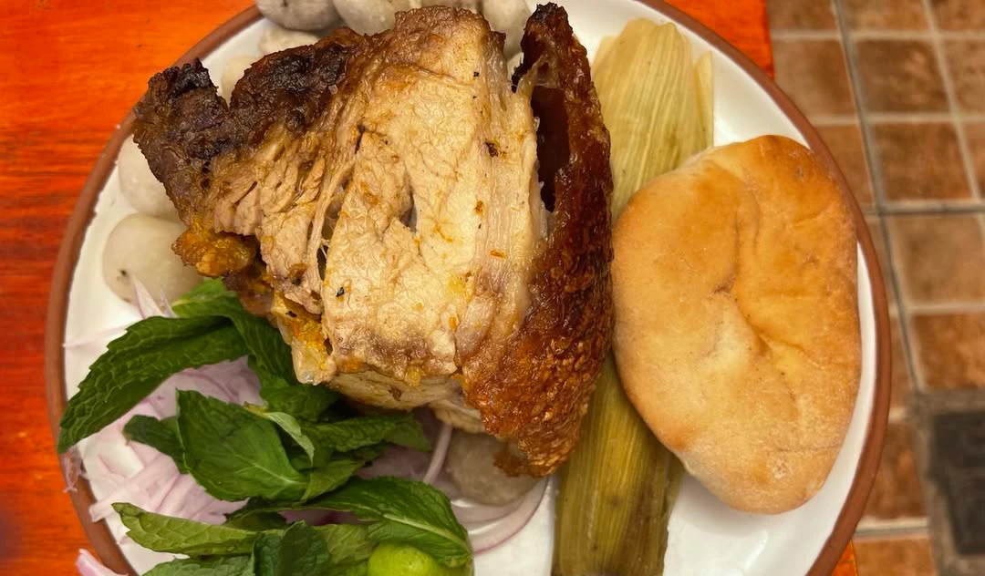 Plate of Cusco-style lechón roast pork with crispy skin, served with tamal, bread, onion slices, mint leaves, and lime.