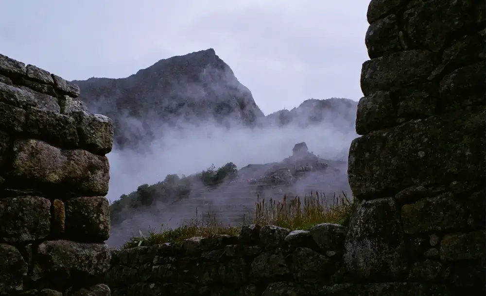 Ruinas de Machu Picchu cubiertas de neblina en un día lluvioso, con terrazas de piedra, montañas verdes, nubes oscuras y una atmósfera mística en los Andes cerca de Cusco.