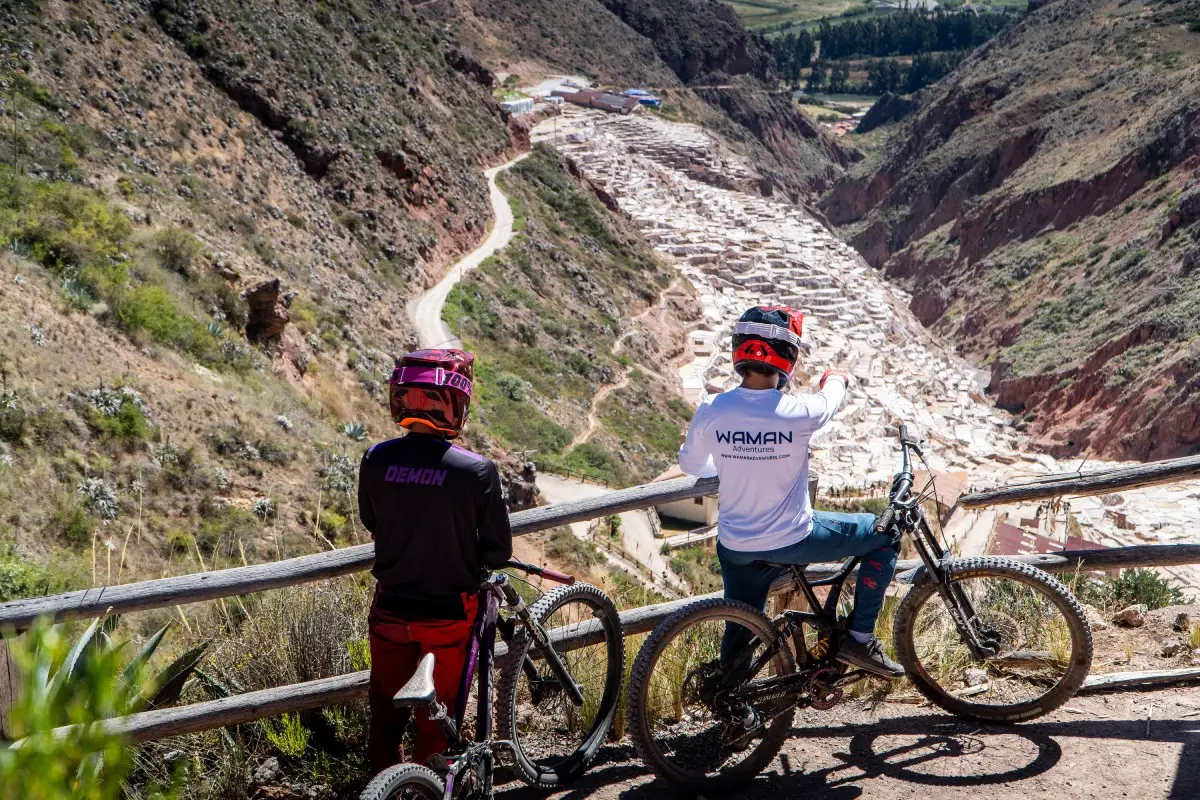 Mountain Biking in Cusco: Two cyclists resting at the Maras Salt Mines viewpoint, with panoramic view of the Sacred Valley, white salt terraces, and reddish mountains (Peru Biking Tours).