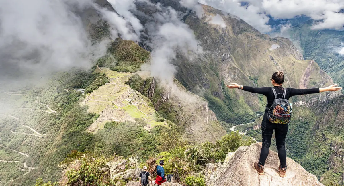 Viajero contemplando Machu Picchu desde un mirador, con terrazas incas, laderas de bosque nuboso y la montaña Huayna Picchu en Cusco, Perú.