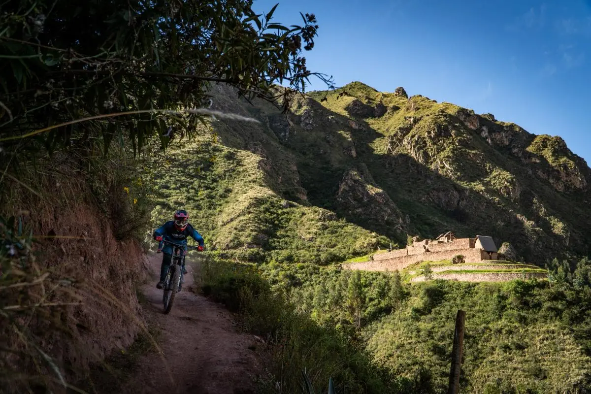 Cyclist descending a forested trail beside Perolniyoc waterfall in the Sacred Valley, surrounded by vegetation, sunlight, and Inca archaeological remains (Peru Biking Tours).