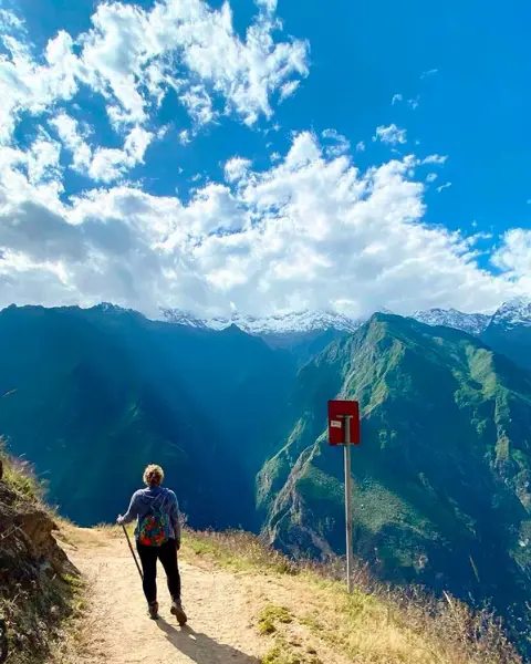 Viajero caminando rumbo a Choquequirao por un sendero de montaña cerca de Cusco, Perú, con valle profundo, laderas verdes y nubes dispersas.