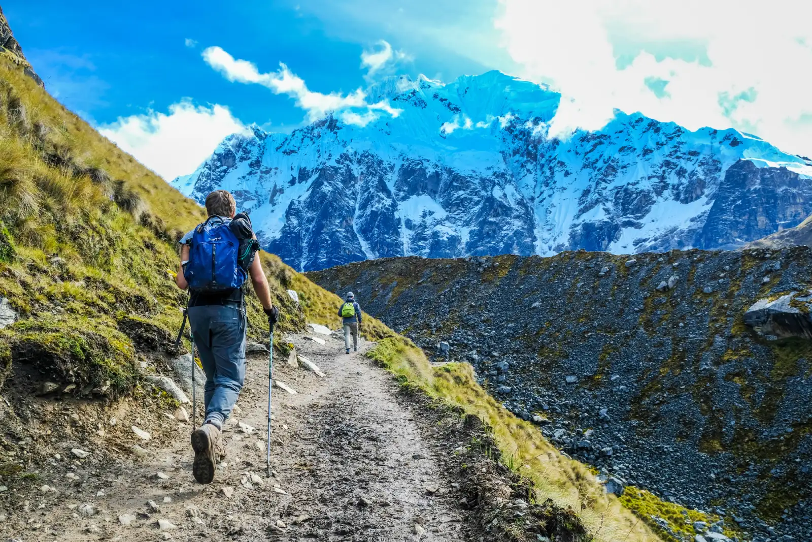 Hiker with trekking poles walking on a high-altitude trail below snow-covered Salkantay Mountain, near the Salkantay Pass section of the trek.