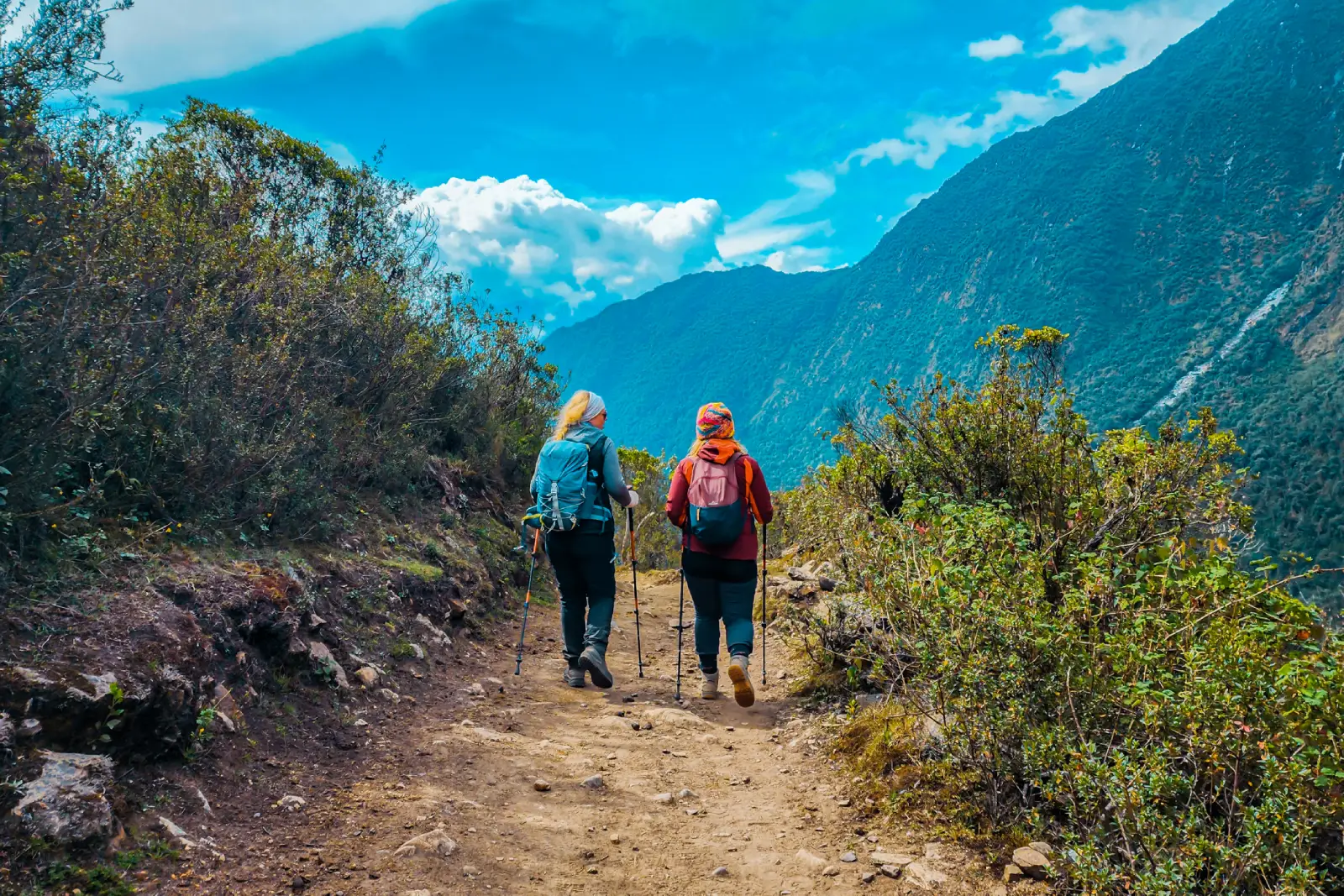 Two hikers walking with trekking poles on a scenic Salkantay Trek trail, surrounded by Andean vegetation and mountain valley views.