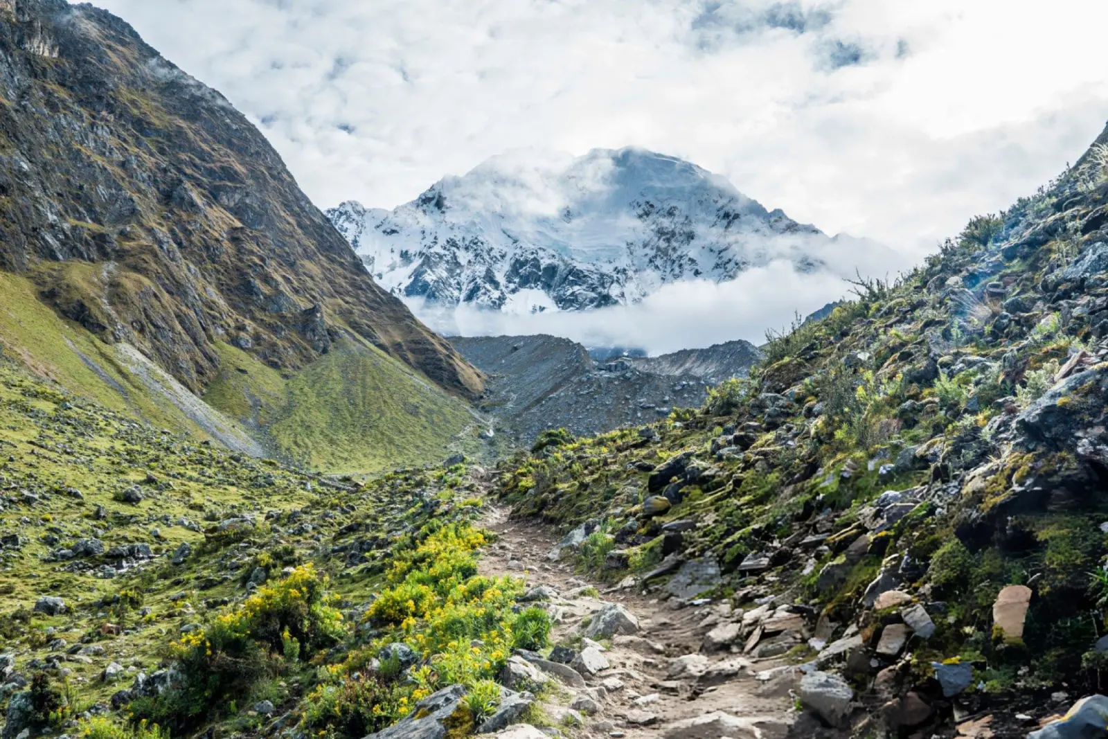 Hiking trail below Salkantay Mountain with green valley slopes and cloud-wrapped glaciers.