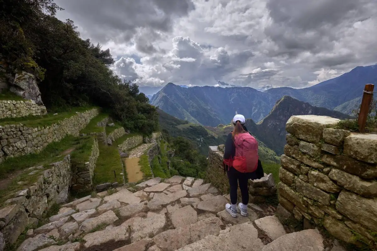 Viajero observando Machu Picchu desde las escalinatas de piedra del Intipunku, con el Huayna Picchu y el valle del Urubamba al fondo.