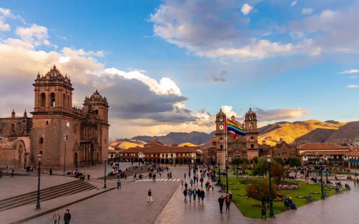 Vista amplia de la Plaza de Armas en Cusco, Perú, con la Catedral del Cusco, edificios coloniales, gente caminando y montañas bajo cielo brillante.