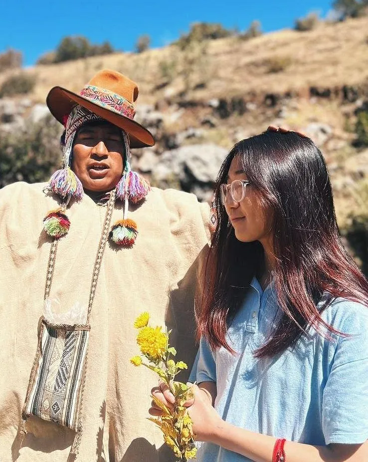 Fotografía de chamán andino (paqo) vestido con poncho tradicional, sombrero decorado y ojotas, guiando ceremonia de Wachuma a participante que sostiene flores amarillas, en paisaje montañoso del altiplano bajo cielo azul
