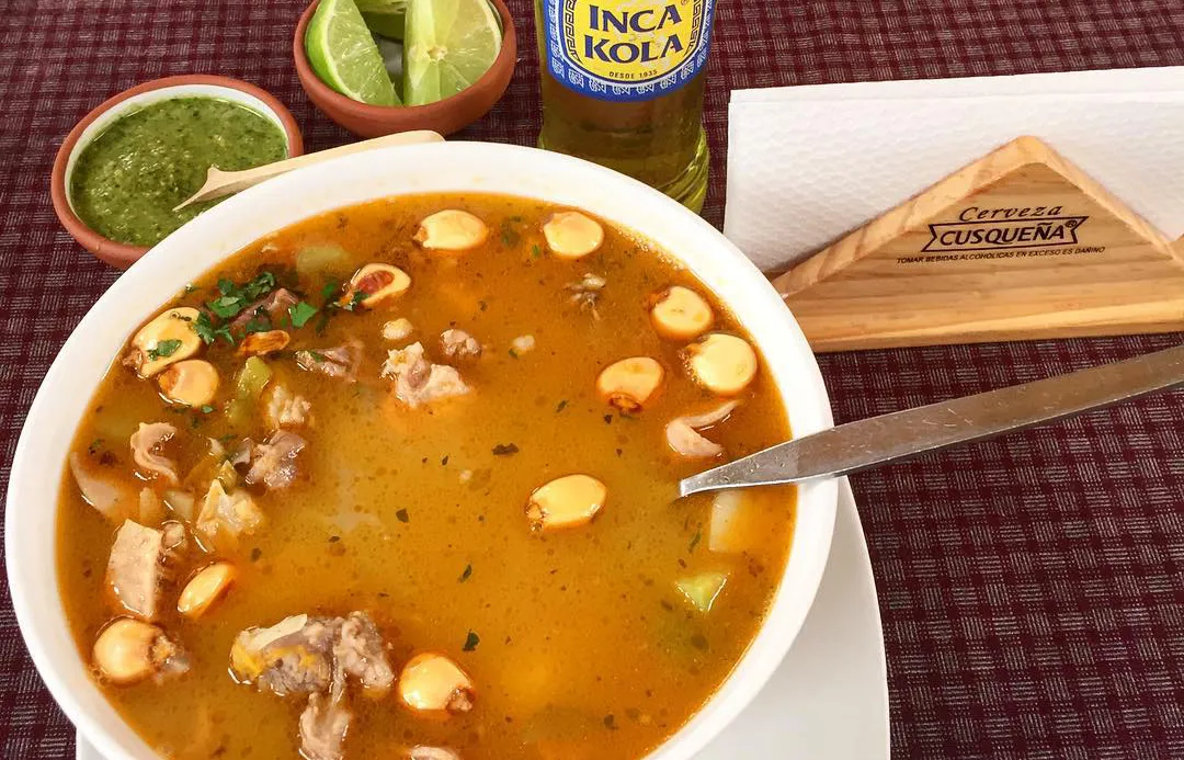 Bowl of Andean chairo soup with meat and vegetables, served with lime wedges, green chili sauce, and a bottle of Inca Kola on the table.