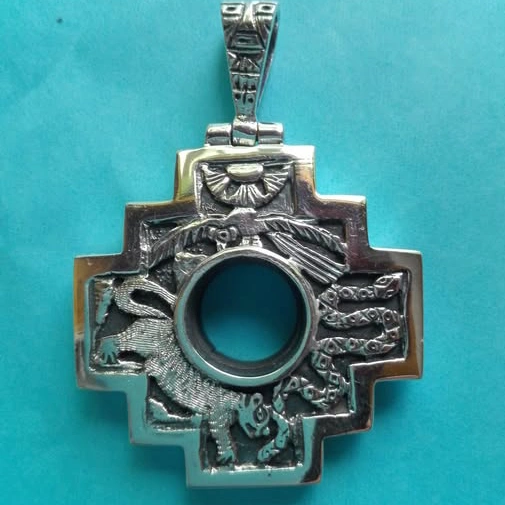 Silver Andean chakana cross pendant with a circular center opening and detailed engraved figures on a turquoise background.