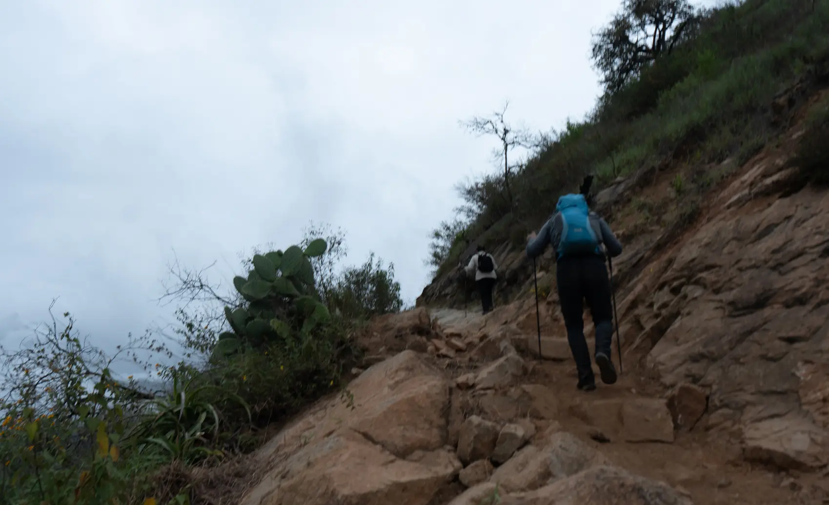 Two hikers climb a steep, rocky Choquequirao Trek path with backpacks, surrounded by green mountains and cloudy Andean sky in the Apurímac valley.