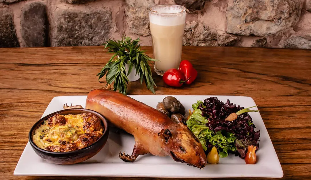 Cusco-style cuy guinea pig al horno served while on a rectangular plate with potatoes, salad, and a baked side dish, with a glass of chicha in the background.