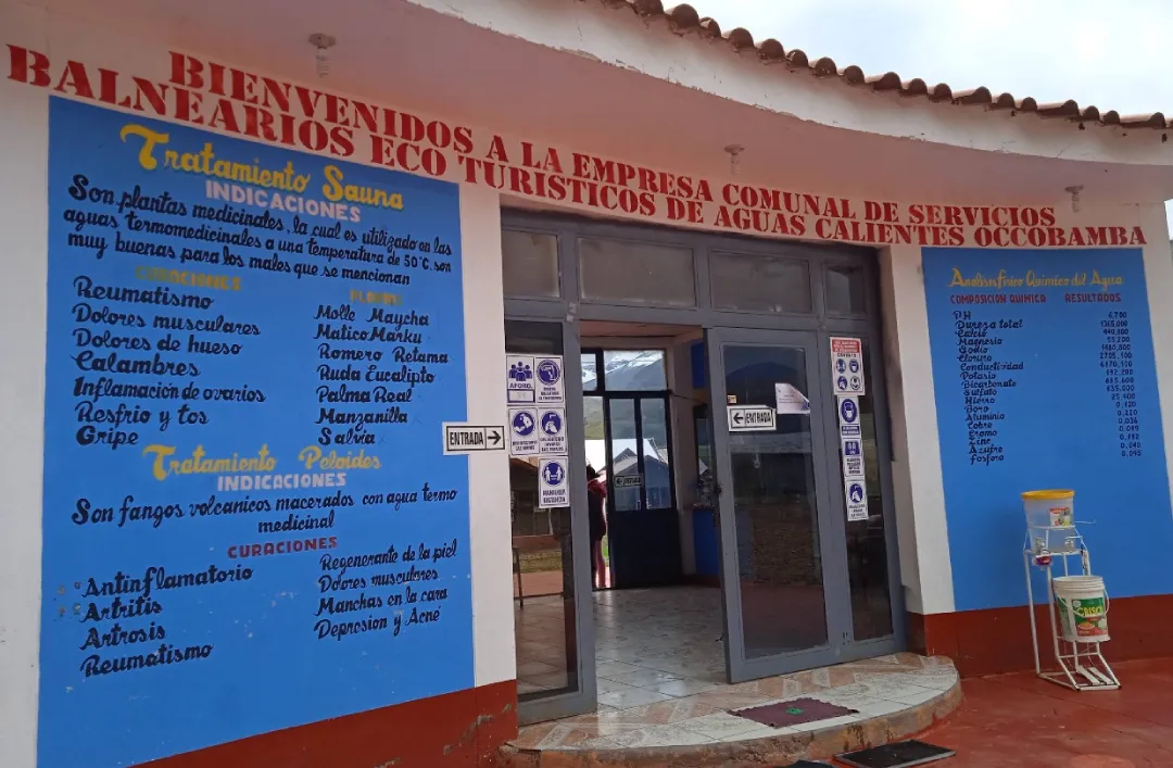Photograph of main entrance to Occobamba Hot Springs Eco-Tourism Spa featuring blue informational murals showing sauna treatments, medicinal indications for rheumatism, muscle pain, and chemical composition of thermal water, access door to center and welcome signage