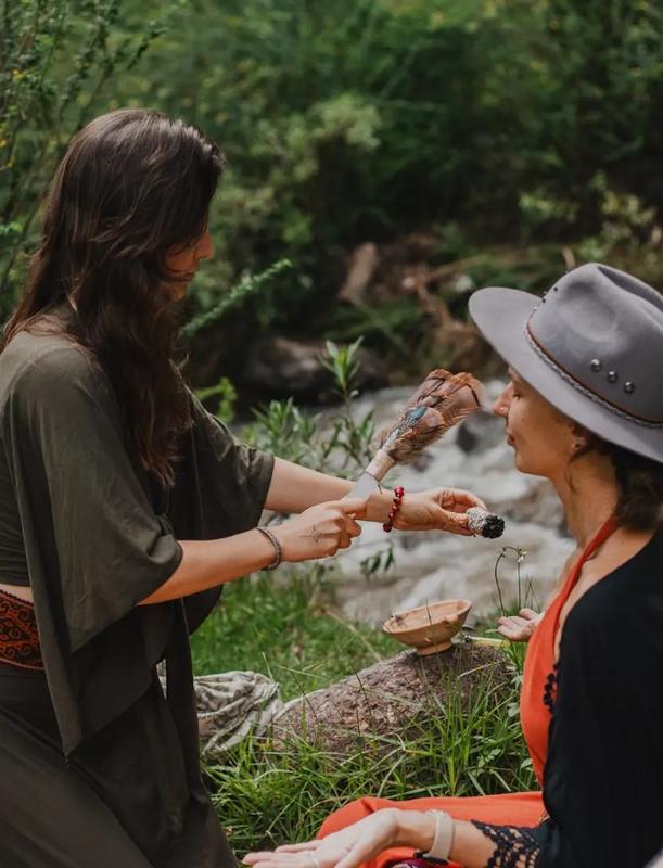 Fotografía de ceremonia de Wachuma con curandera realizando ritual de limpieza energética a participante usando sahumerio y plumas sagradas, en entorno natural junto a río, mostrando práctica ceremonial ancestral andina con San Pedro