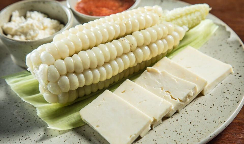 Peruvian giant corn (choclo) served with slices of fresh cheese on a plate, with sauces in small bowls in the background.