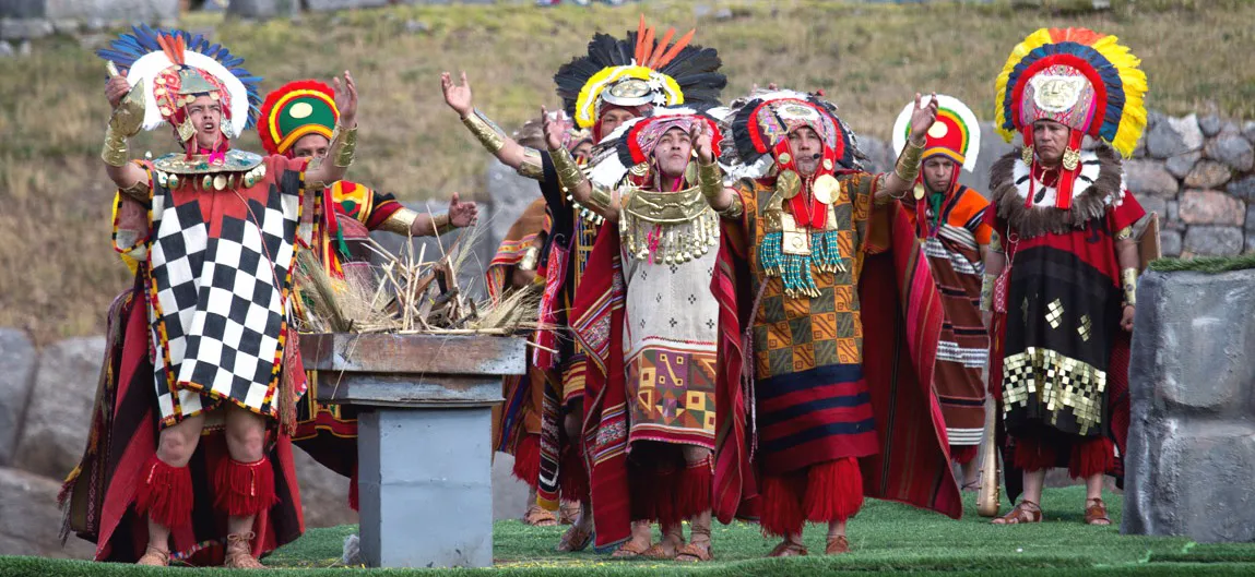 Group of Inti Raymi ceremony performers wearing colorful Inca-style costumes and feathered headdresses, standing around an altar outdoors.
