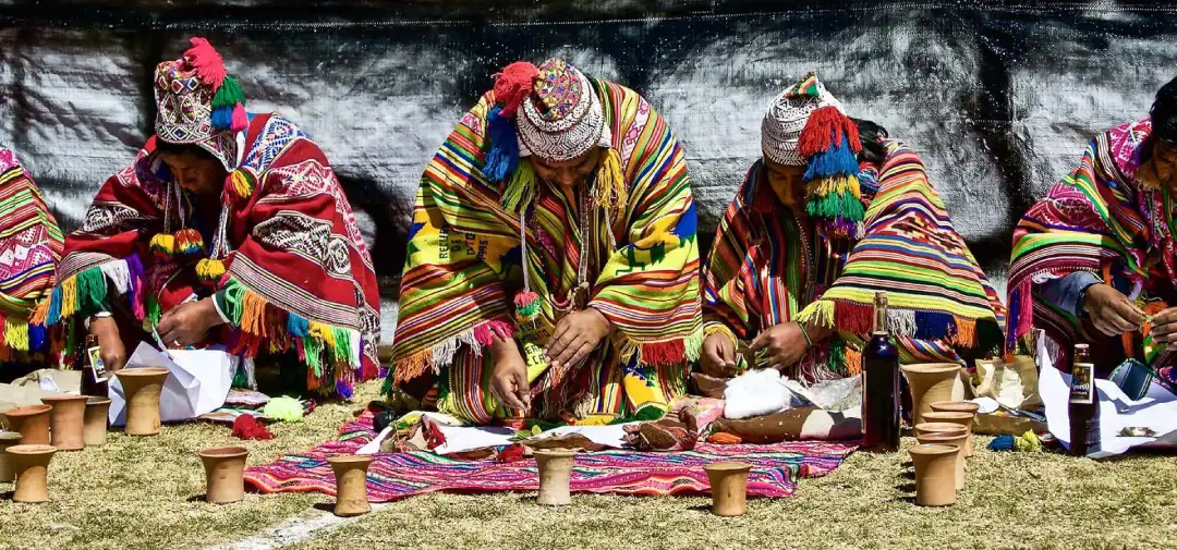 Andean spiritual leaders in colorful traditional clothing preparing a Pachamama offering ceremony with ritual items laid out on woven textiles.