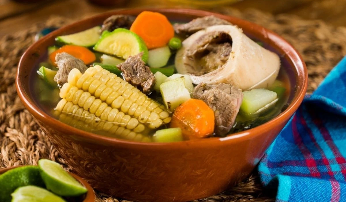 Bowl of Peruvian puchero soup with beef, corn on the cob, squash, carrots, and vegetables in a clear broth.