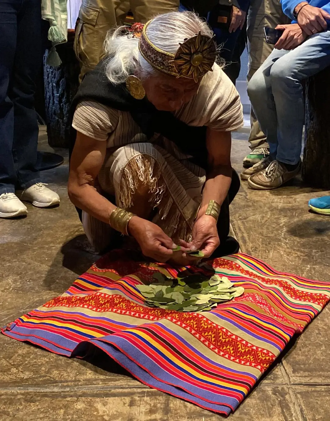 Photograph of elderly Andean woman kneeling performing coca leaf reading ceremony on multicolored aguayo, dressed in traditional clothing and ceremonial headdress, surrounded by people observing the ancestral ritual