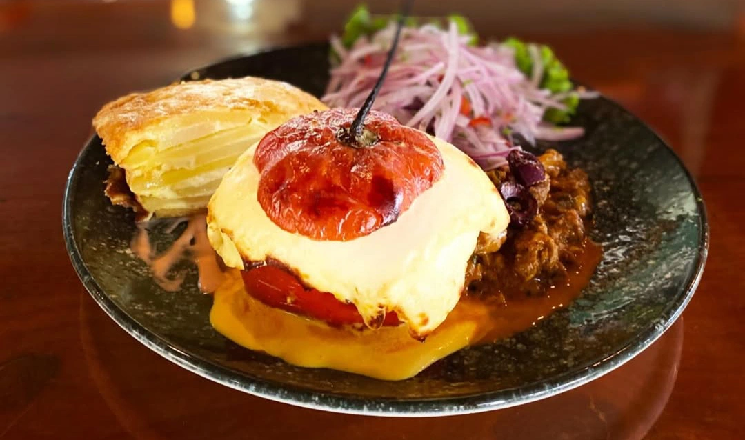 Stuffed rocoto pepper topped with melted cheese, served with pastel de papa (potato casserole) and a red onion salad on a black plate.