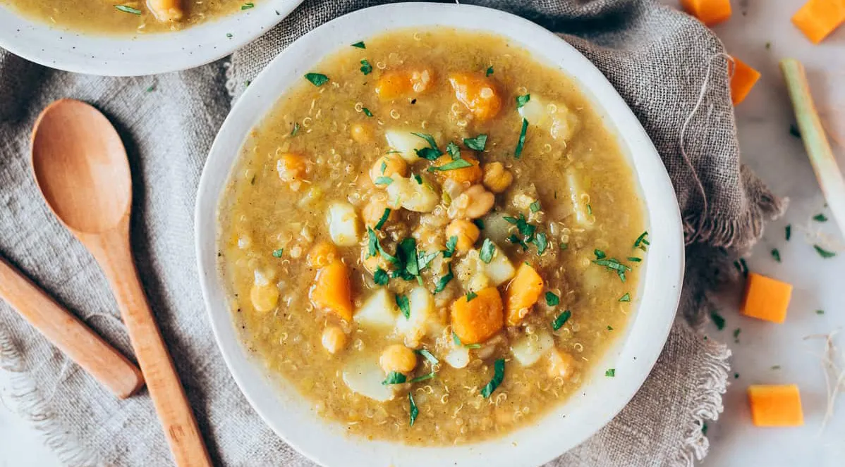 Bowl of quinoa soup with chickpeas, potatoes, carrots, and herbs, served on a rustic cloth with a wooden spoon nearby