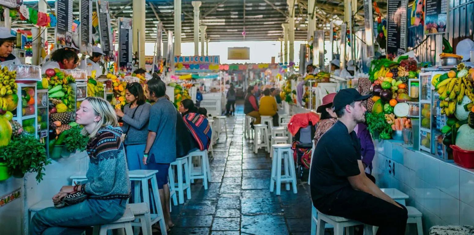 Interior photograph of San Pedro Market in Cusco showing central aisle with fresh juice and fruit stalls, tourists and locals sitting on white benches tasting beverages, colorful hanging decorations, abundant bananas, pineapples and tropical fruits on display